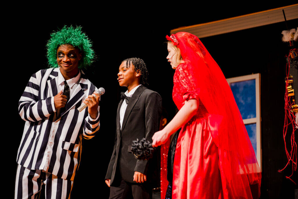 Three people in costume perform on stage. One wears a green wig, striped suit, and holds a mic; another wears a black tuxedo; the third wears a red dress and veil, holding a black bouquet in front of a set with a painted window.