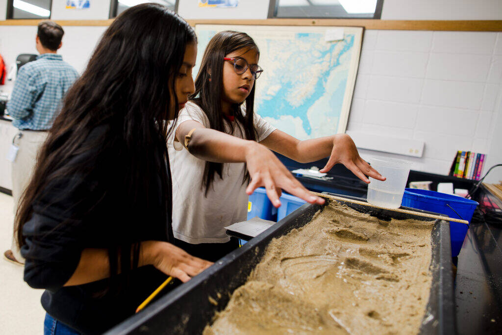 Two students conduct a science experiment with wet sand in a classroom, using their hands to shape the surface. A map is on the wall in the background.